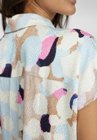 Chemise à manches courtes en tissu léger présentant un motif abstrait multicolore dans des tons pastel de bleu, rose et marron.