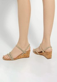 Sandalen met een sleufhak van kurk, versierd met kruislings lopende metalen banden. Kleur: lichtgoud. Open teenontwerp onthult de tenen.