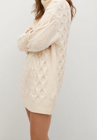 Vaalea beige kaapelikudottu neulepaita, jossa on korkea kaulus, monimutkaisia kuvioita ja ribattuja hihansuita, pidentyy keskivartalon pituuteen.