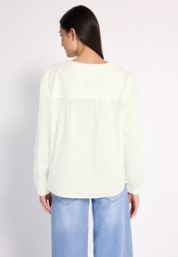 Camicia bianca a maniche lunghe con scollatura rotonda e schiena plissettata. Abbinata a jeans a gamba larga di colore blu chiaro, che mostrano una vestibilità rilassata e una consistenza morbida.
