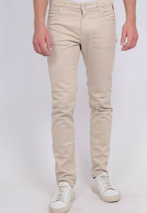 Homme portant un pantalon beige coupe slim et des baskets blanches à lacets, debout devant un fond clair uni.