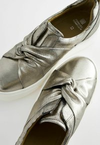 Metallic silver sneakers with twisted fabric design, elastic entry, cushioned sole, and white rubber outsole. Smooth texture and sleek appearance.