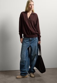 Pull en laine marron foncé, oversize, avec un col en V, associé à un jean bleu clair, à jambes larges. Chaussures décontractées beiges, tenant un grand sac fourre-tout noir.