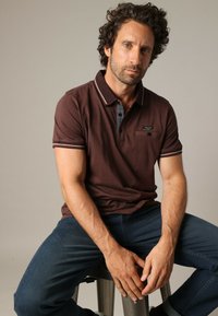 Polo shirt marron avec col rayé contrastant, patte de boutonnage et poche poitrine. Le modèle porte un jean bleu et est assis sur un tabouret.