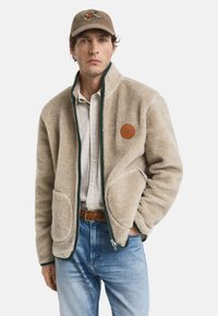 Uomo che indossa una giacca in pile beige sopra una camicia a quadri, jeans blu, una cintura intrecciata marrone e un cappello beige con ricamo di un uccello.