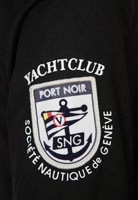 Tessuto nero con un distintivo ricamato bianco che presenta un design a scudo, un'ancora e testo: "Yachtclub Port Noir Société Nautique de Genève."