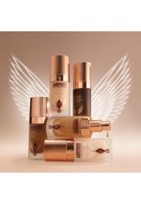 Five foundation bottles with gold accents, varying in shade. One bottle has a pump, while others have screw-top lids. Neutral background with wing design.