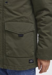 Chaqueta de tela pesada y verde que presenta dos bolsillos con solapa, acentos negros y una etiqueta de marca visible en el dobladillo. Textura suave, diseño casual.