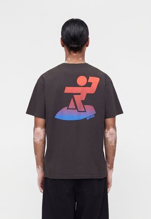Personne aux cheveux foncés en chignon portant un t-shirt noir avec un graphisme stylisé de surfeur rouge et bleu dans le dos.