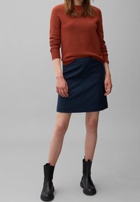 Rostfarbener Pullover, marineblauer Rock mit einer Seitentasche und schwarze Stiefeletten. Der Stoff ist strukturiert, mit einem figurbetonten Schnitt und minimalen Details.
