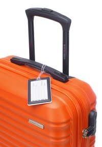 Valise rigide orange avec texture striée verticale, poignée noire rétractable et étiquette pour informations personnelles attachée.