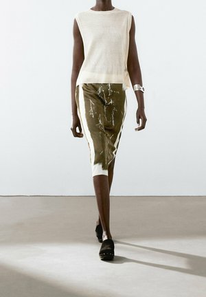 Mannequin marchant dans un haut blanc sans manches, une jupe midi olive avec des rayures verticales blanches, des chaussures noires cloutées et un bracelet manchette argenté.