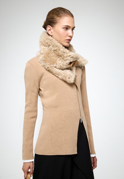 Donna che indossa un maglione beige con zip e un ampio colletto morbido, una gonna nera, mentre tiene una borsa beige su uno sfondo semplice.