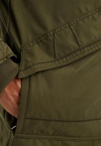 Oljnozelena jakna s glatkom teksturom, s istaknutim šavovima, rebrastim manžetama i bočnom džepom koji se vidi na donjoj lijevoj strani.