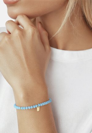 Femme portant un bracelet bleu clair à perles avec un petit charm rectangulaire doré, reposant sa main près du menton, vêtue d'une chemise blanche.
