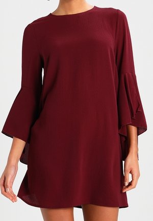 Kobieta w bordowej sukience do kolan z długimi rękawami, rozkloszowanymi, warstwowymi rękawami i okrągłym dekoltem.