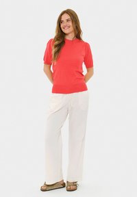 Maglione corto a maniche corte color corallo con un design aderente, abbinato a pantaloni bianchi larghi e sandali marrone chiaro con dettagli a fiocco.