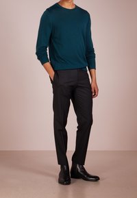 Teal, long-sleeve sweater paired with black, slim-fit trousers and black leather ankle boots. Model stands with one hand in pocket.