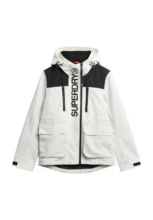 Veste blanche et noire avec capuche, grandes poches avant, deux poches à fermeture éclair sur la poitrine, et logo « SUPERDRY » bien visible sur le devant.