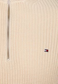 Pull en maille gaufrée de couleur crème avec une fermeture éclair verticale en argent et un petit logo de drapeau rectangulaire rouge, blanc et marine sur la poitrine.