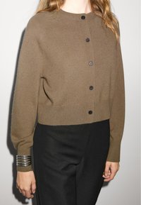 Brown knitted cardigan with black buttons, round neckline, and long sleeves. Worn over dark trousers, with a silver bracelet on the wrist.