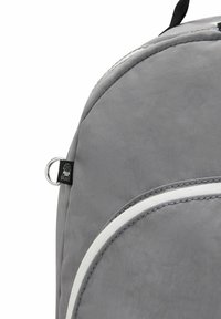 Sac à dos gris avec des accents blancs, fabriqué en tissu résistant ; il dispose d'une fermeture éclair, d'une boucle en métal circulaire et d'une étiquette de logo.
