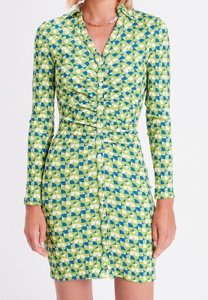 Femme portant une robe courte à manches longues boutonnée avec un motif géométrique vert, bleu et blanc et un collier chaîne en or.