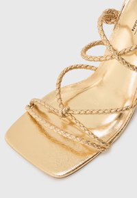 Sandalias doradas con tiras trenzadas delgadas, un acabado metálico brillante y un diseño de punta cuadrada. Textura suave y estética minimalista.