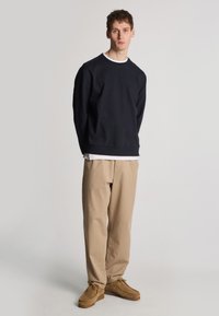 Felpa blu con polsini e orlo a coste, indossata sopra una camicia bianca. I chinos beige e le scarpe marroni completano il look, caratterizzato da una vestibilità rilassata.