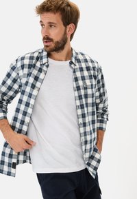 Męska koszula zapinana na guziki w niebiesko-białą kratę, z kieszenią, nałożona na biały t-shirt. Materiał wygląda na bawełnę, a koszula ma luźny krój.