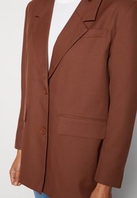 Mujer vistiendo un blazer marrón con botones y bolsillos de solapa sobre una blusa blanca y jeans azules, mostrada de los hombros a las caderas.