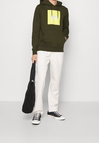 Sudadera verde oliva con un gráfico cuadrado amarillo, pantalones blancos, bolsa tote negra y zapatillas negras con suelas blancas. Atuendo casual.