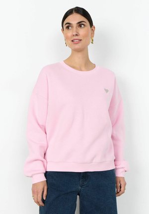 Woman wearing a light pink long-sleeve sweatshirt and dark blue jeans, standing against a plain white background, looking slightly right.
