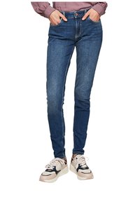 Štíhlé modré džíny z denimu s pěti kapsami, knoflíkem a zipem, nošené s světlými, mohutnými teniskami.