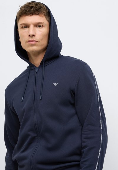 Σκούρο μπλε φούτερ με φερμουάρ, κατασκευασμένο από απαλό ύφασμα, με κουκούλα που σφίγγεται με κορδόνι και λογότυπο στο στήθος, με κείμενο κατά μήκος του μανικιού.