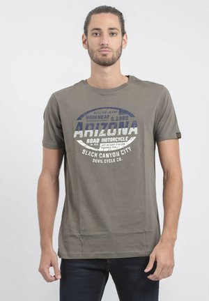 T-shirt en coton vert olive avec des manches courtes, un col rond, présentant un texte graphique et un logo en blanc et bleu. Design simple et droit.