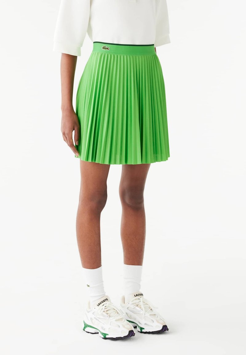Zielona plisowana spódnica z dopasowanym pasem, z małym akcentem logo. Noszona z białymi skarpetkami i trampkami. Gładka faktura.