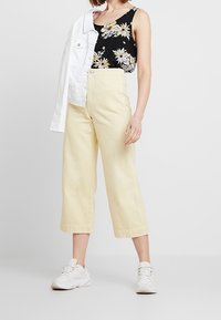 Top in tessuto nero con fiori gialli, abbinato a pantaloni culotte gialli a vita alta e una giacca bianca. Sneakers bianche completano il look.