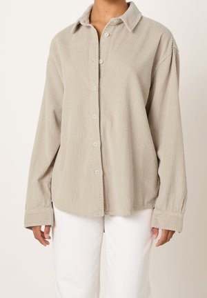 Personne portant une chemise en velours côtelé beige boutonnée et un pantalon blanc, debout devant un fond clair uni.