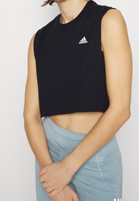 Černý bezrukávový crop top z bavlny s malým bílým logem Adidas na hrudi. V kombinaci s světle modrými sportovními legínami.