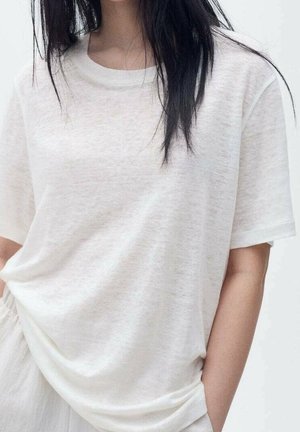 Mujer con cabello largo y oscuro que lleva una camiseta blanca lisa y holgada de manga corta y con una mano en el bolsillo.