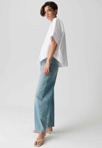 Vrouw draagt een losse witte top met korte mouwen, lichtblauwe jeans met wijd uitlopende pijpen en beige sandalen met hak, staand tegen een effen lichte achtergrond.