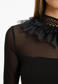 Schwarzes Oberteil mit durchscheinendem Mesh-Overlay und bogenförmigem Kragen. Der Kragen ist mit strukturiertem schwarzem Netzstoff und satinähnlichen Rüschen verziert.