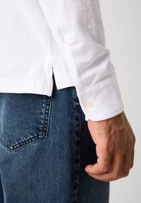 Die Hand eines Mannes ruht nahe der Gesäßtasche einer blauen Jeans, während er ein weißes Langarmshirt mit geknöpften Manschetten und seitlichem Schlitz trägt.