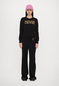 Melna džemperis ar zelta "Versace Jeans Couture" logo, apvienots ar plašām melnām biksēm. Izskatu papildina rozā cepure ar akcentiem.