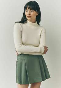 Cremefarvet ribbet turtleneck sweater parret med en grøn plisseret nederdel. Sweateren har lange ærmer, og nederdelen har skarpe pliser.