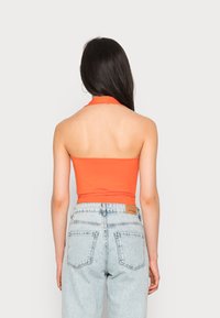 Top alaranjado de decote halter feito de tecido stretch, combinado com jeans de cintura alta azul claro com um patch de logo em couro e costuras visíveis.