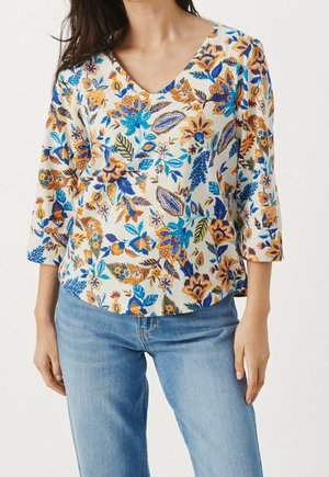 Femme portant un chemisier blanc à col en V avec des motifs floraux bleus, jaunes et oranges et un jean bleu clair sur un fond uni.