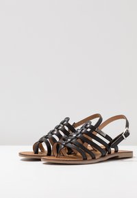 Sandalias negras con tiras, de diseño abierto, que cuentan con una suela plana y un cierre de hebilla. El material es suave y con una textura trenzada.