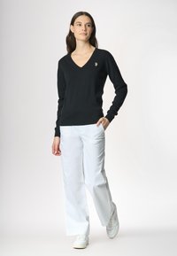 Donna con i capelli lunghi che indossa un maglione nero a V, pantaloni bianchi larghi e sneakers, in posa contro uno sfondo chiaro.